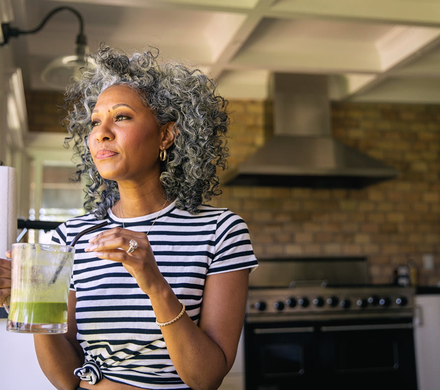A middle-aged woman drinking a green juice out of a class, looking out the window.