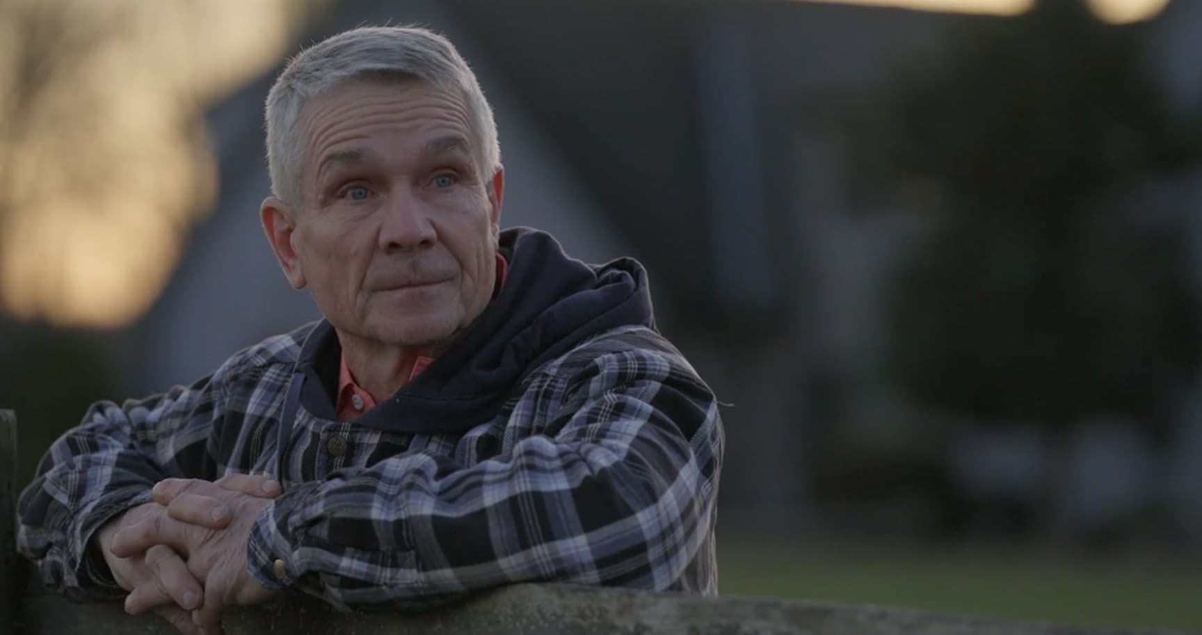 A gray-haired man in a sweater with his arms folded on a fence